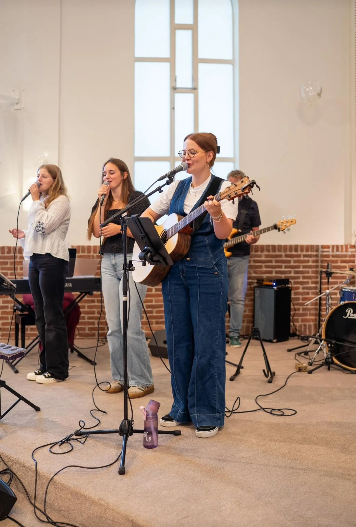 Vrouw met bril die gitaar speelt en zingt, met drie vrouwen en een man die ook zingen en muziek maken, op een podium in een zaal met witte muren en een groot raam, decoratief met ballonnen.