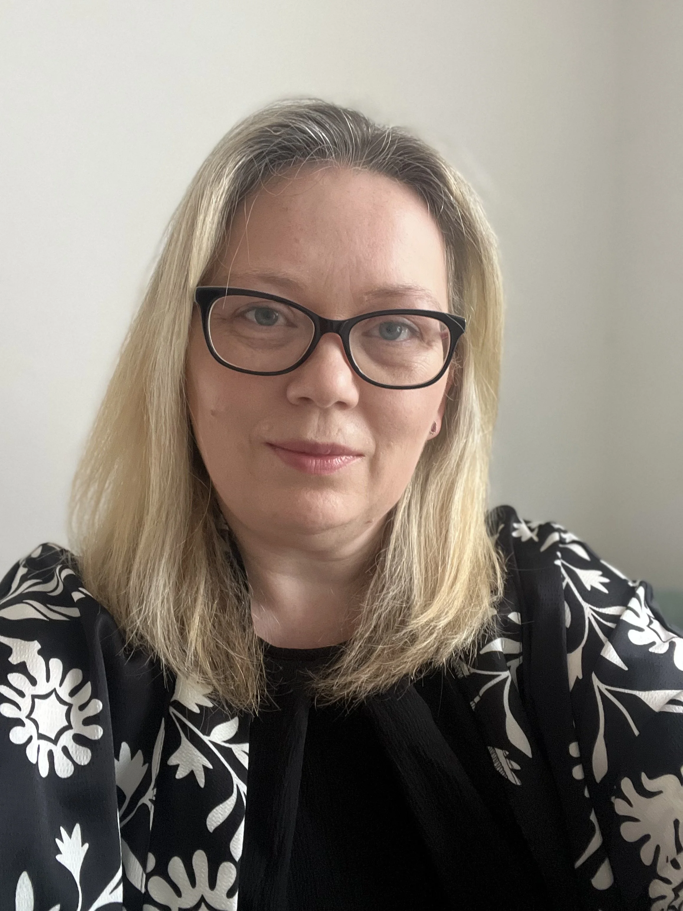 A woman with blonde hair, glasses, and a black and white floral patterned top, taking a selfie indoors against a plain light-colored wall.