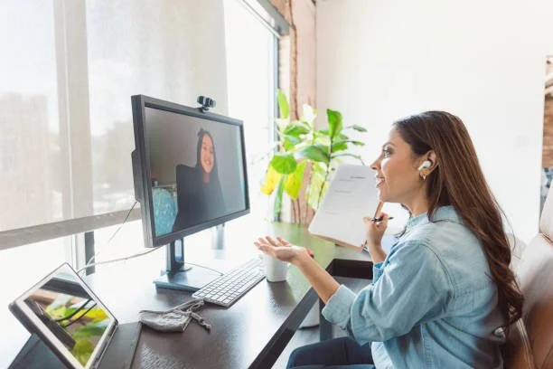 Woman participating in a video call, holding a notebook, sitting at a desk with a monitor, tablet, and plants in a bright office.