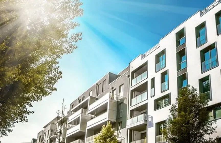 Modern residential apartment buildings with balconies, trees in the foreground, and a clear blue sky with sunlight