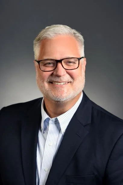 Business professional man with gray hair and beard, wearing glasses, a dark suit, and a light collared shirt, smiling against a dark gray background.