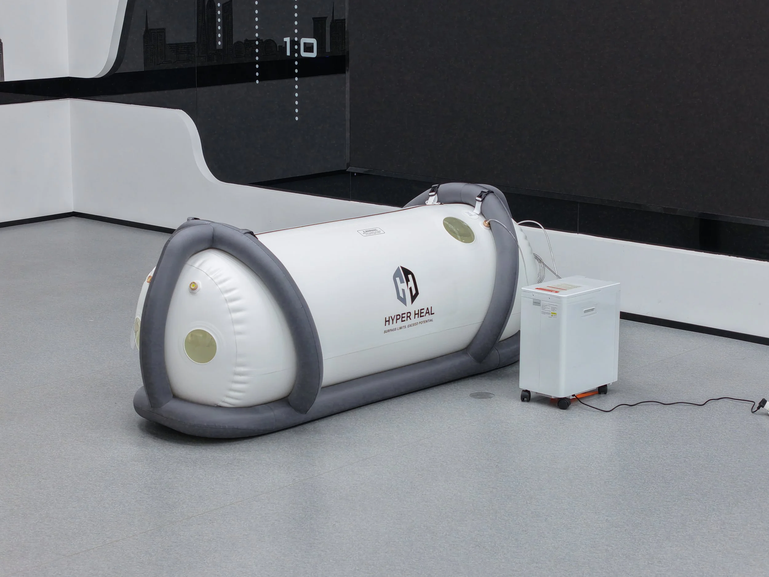 A hyperbaric oxygen therapy chamber lying on the floor, with a white exterior, black padded edges, and the logo 'HYPER HEAL' on it. There is a white machine connected to it on wheels, with cords attached.