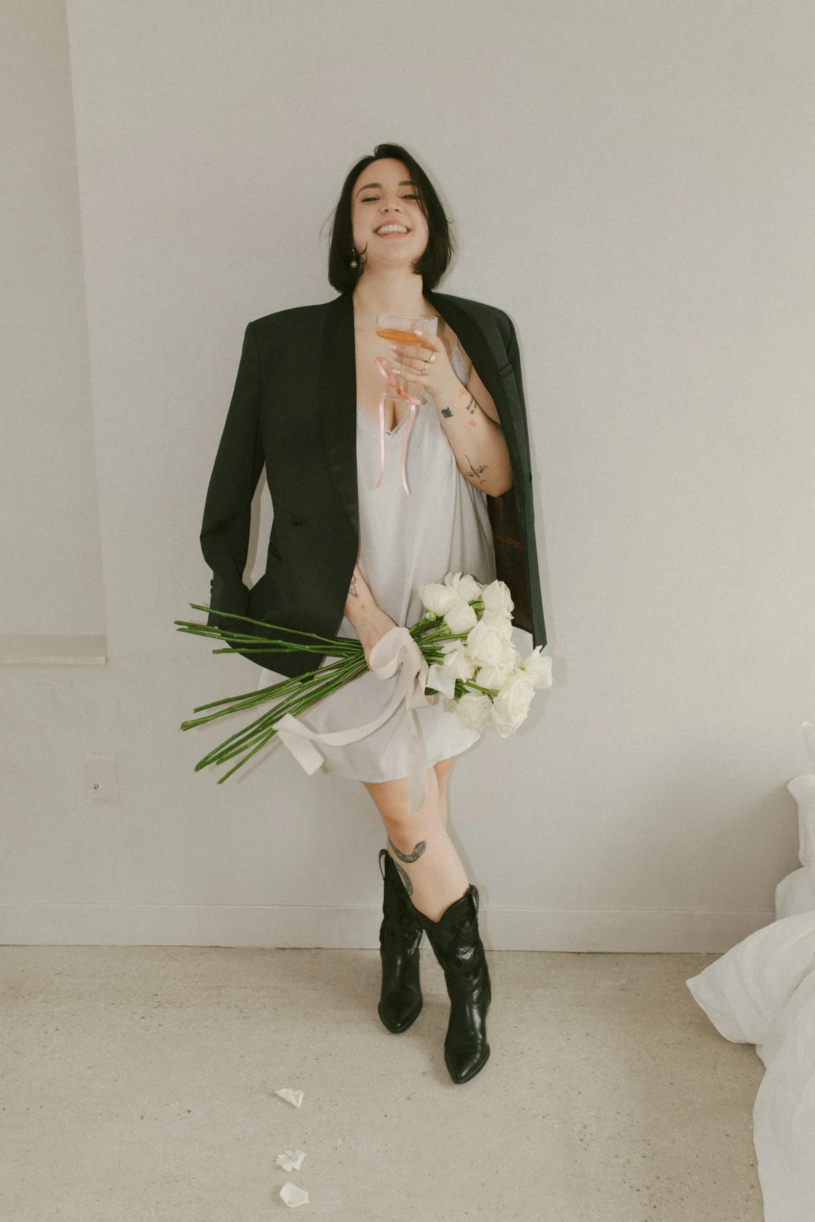 A woman with black hair, wearing a black blazer over a gray dress, holding a bouquet of white roses and a glass of champagne, standing against a plain white wall and smiling.