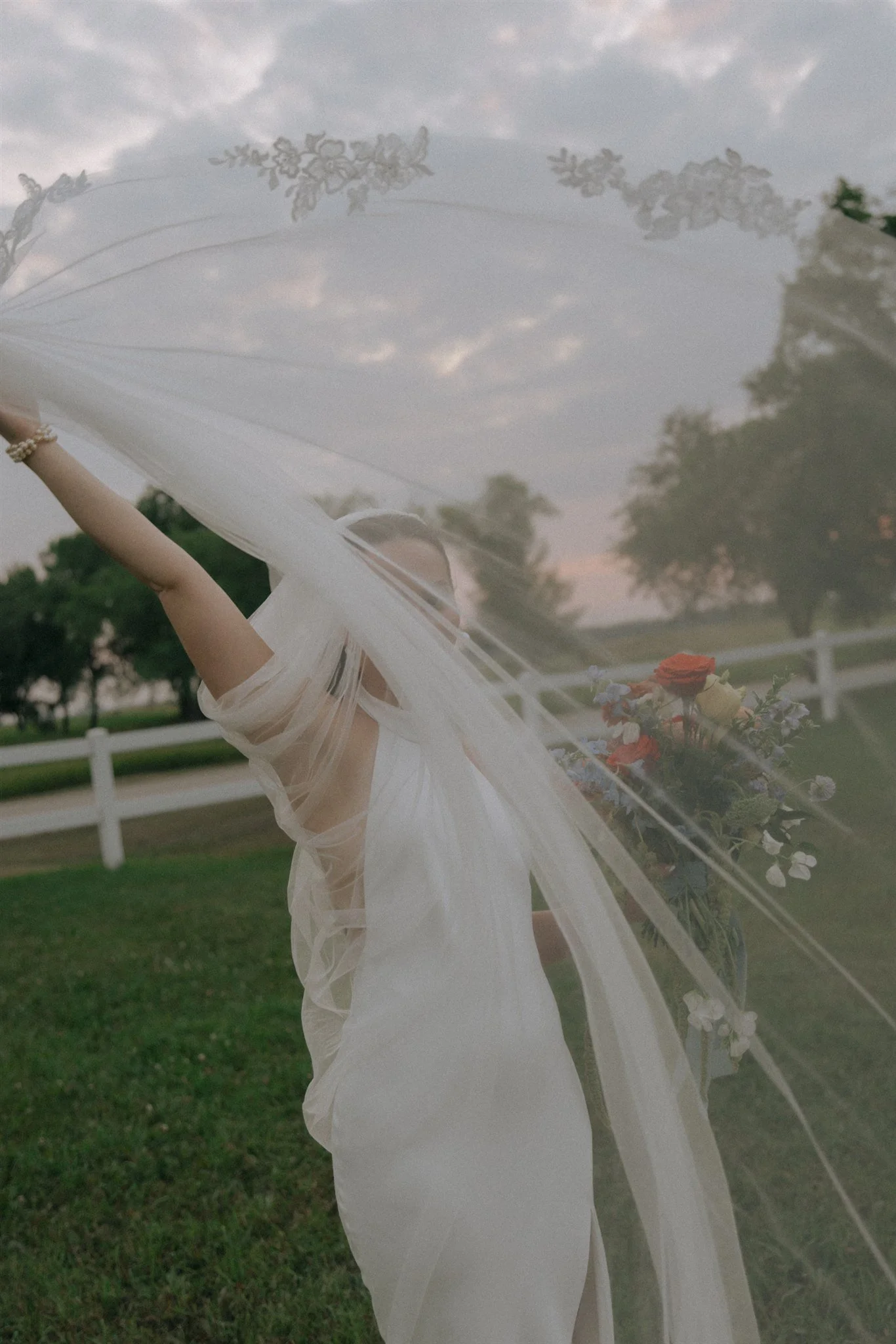 A woman dressed in white, holding a bouquet of flowers, is standing outdoors near a white fence with trees in the background during the evening, seen through a semi-transparent curtain.