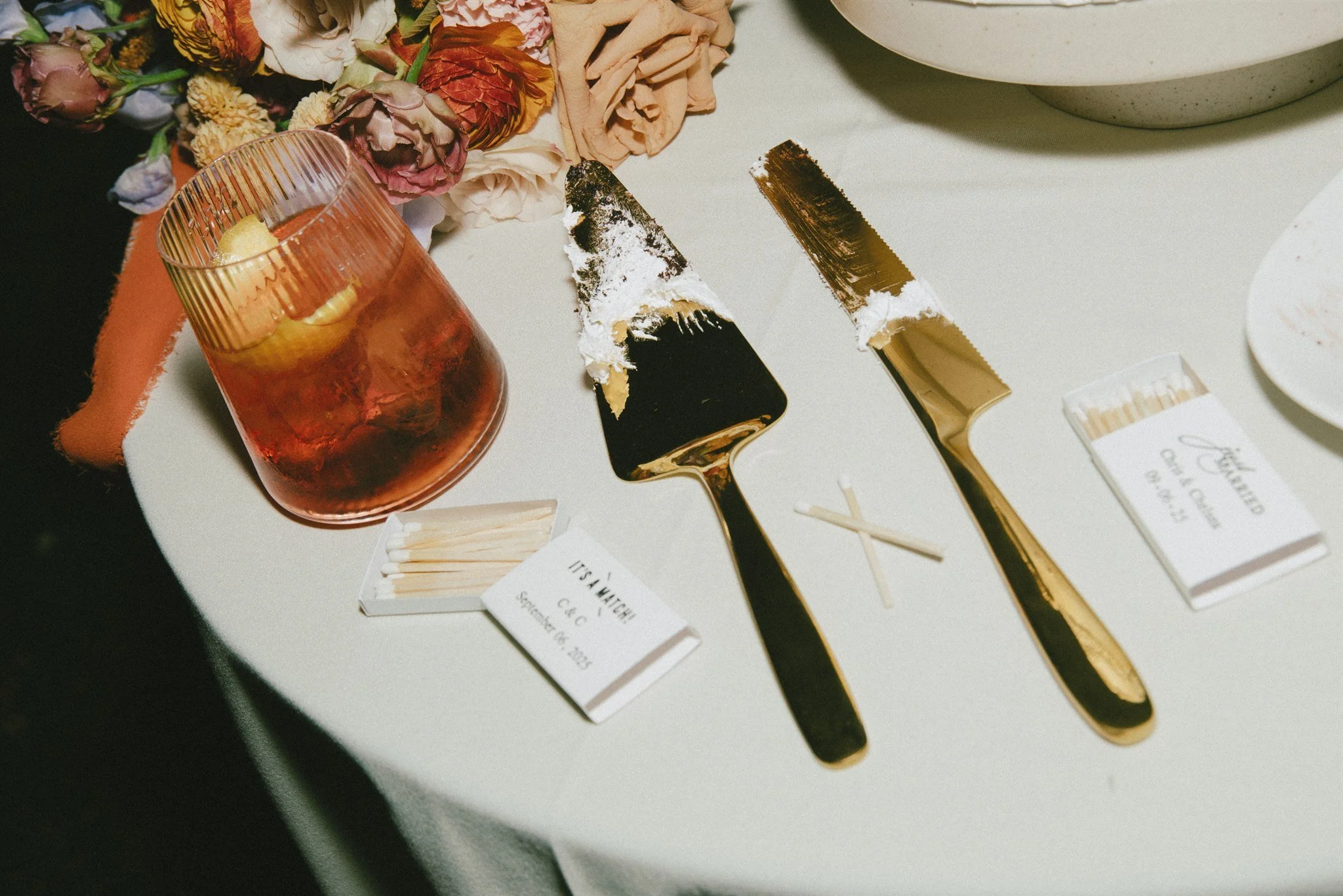 A table with a pink drink containing lemon slices, two burnt-out candles, and gold and black knives with partial white wax residue on the blades. There are flower petals and matchbooks nearby.