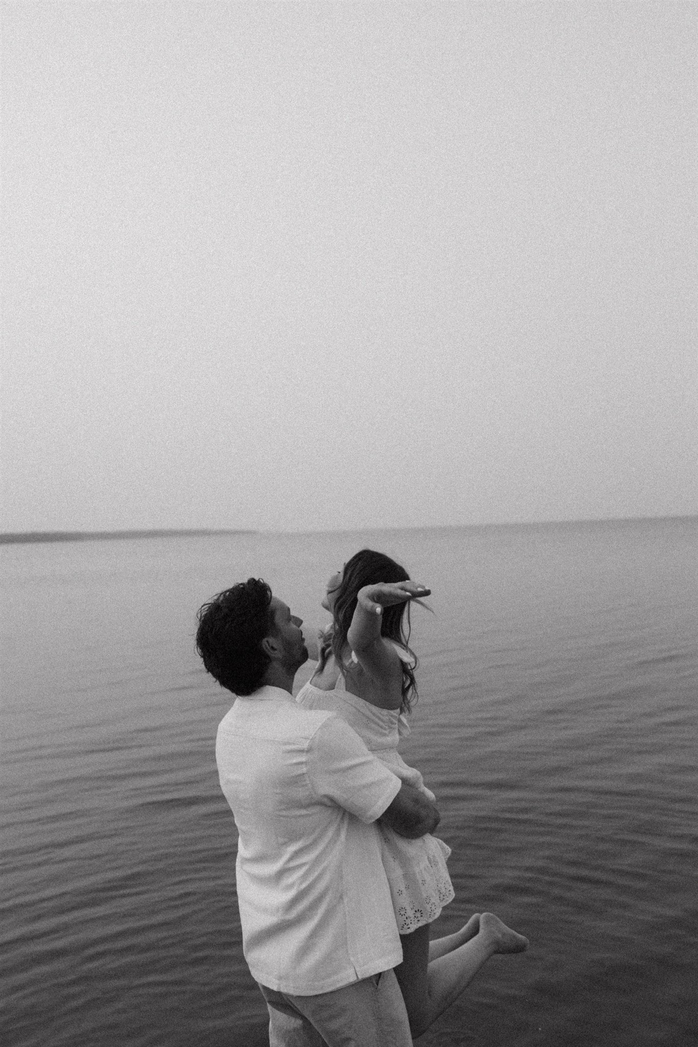 A man holding a woman sideways at the edge of a body of water, with her arm outstretched and barefoot, in black and white.
