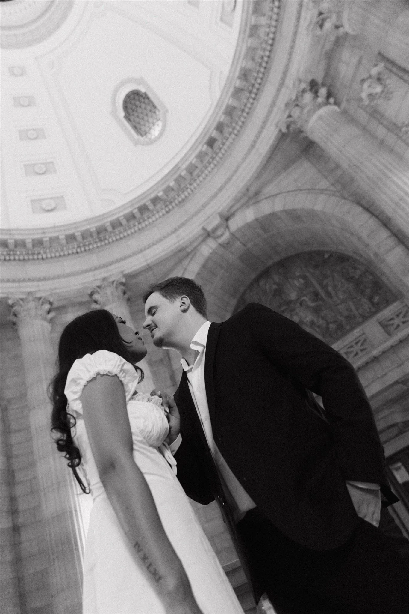 A black and white photo of a couple standing close together, facing each other, inside a grand historic building with ornate columns and a domed ceiling.
