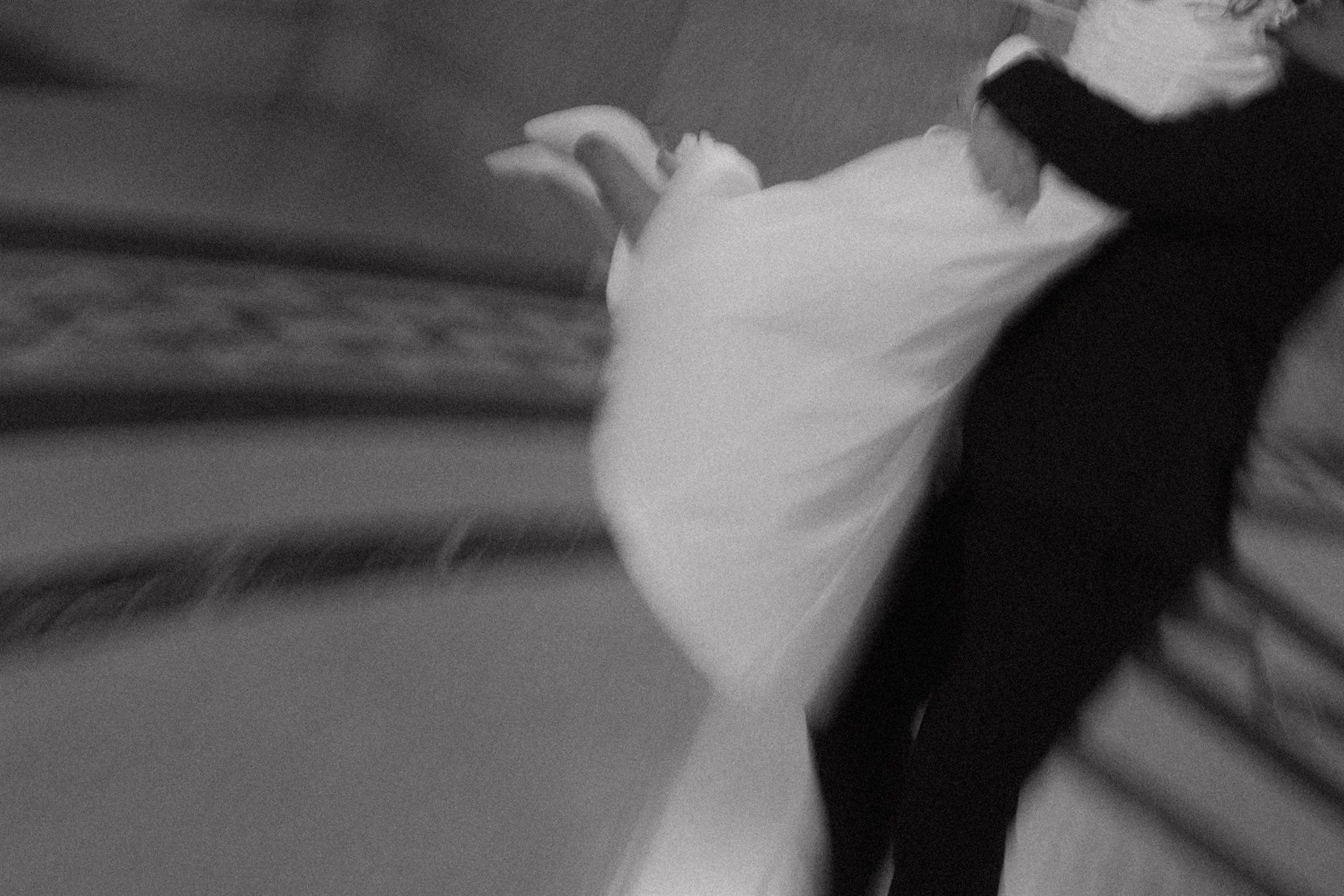 A person lying on a bed with their legs elevated, wearing dark pants and a light-colored top, with a blanket partially covering them. The room has a carpeted floor and a piece of furniture is visible in the background.