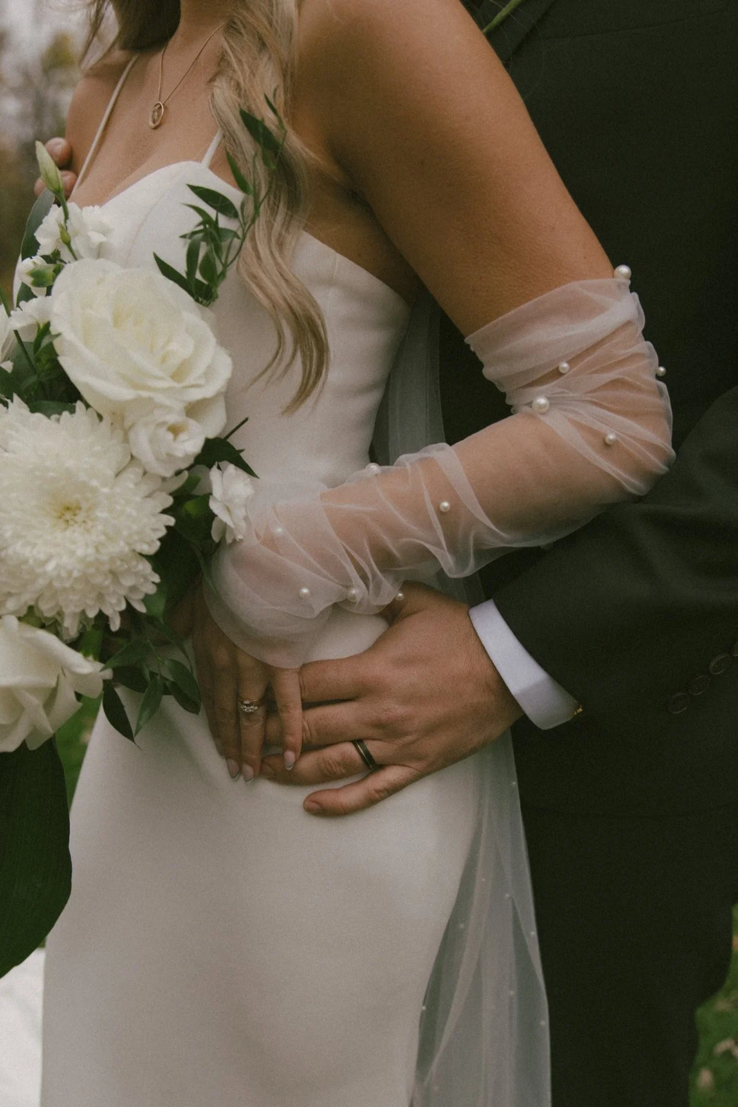 Close-up of a bride and groom holding hands during a wedding, with the bride holding a bouquet of white roses and chrysanthemums, and both wearing wedding rings. The bride is in a white wedding dress with sheer, pearl-embellished sleeves, and the groom in a black suit with a white shirt.