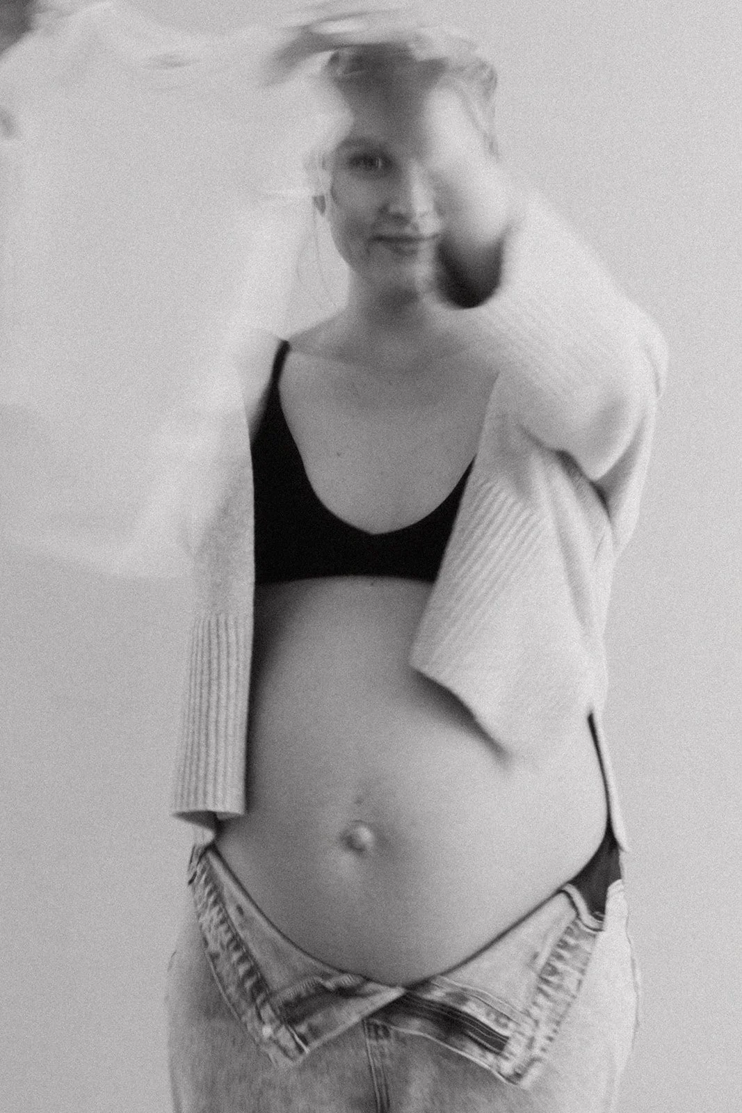 A woman smiling at the camera, wearing a black bra, partially unbuttoned shirt, and jeans, standing against a plain background.