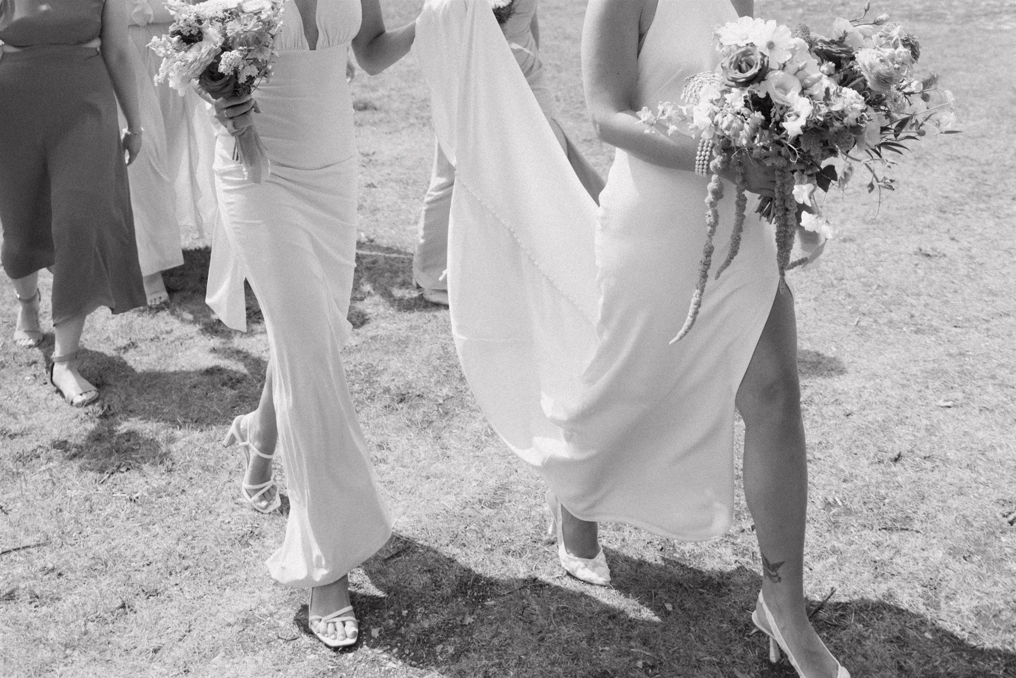 Women wearing white dresses and high heels, holding bouquets of flowers, walking on a grass field.