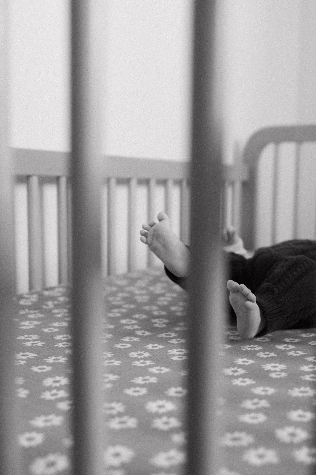 Child lying on a bed with patterned sheets viewed through the bars of a crib.