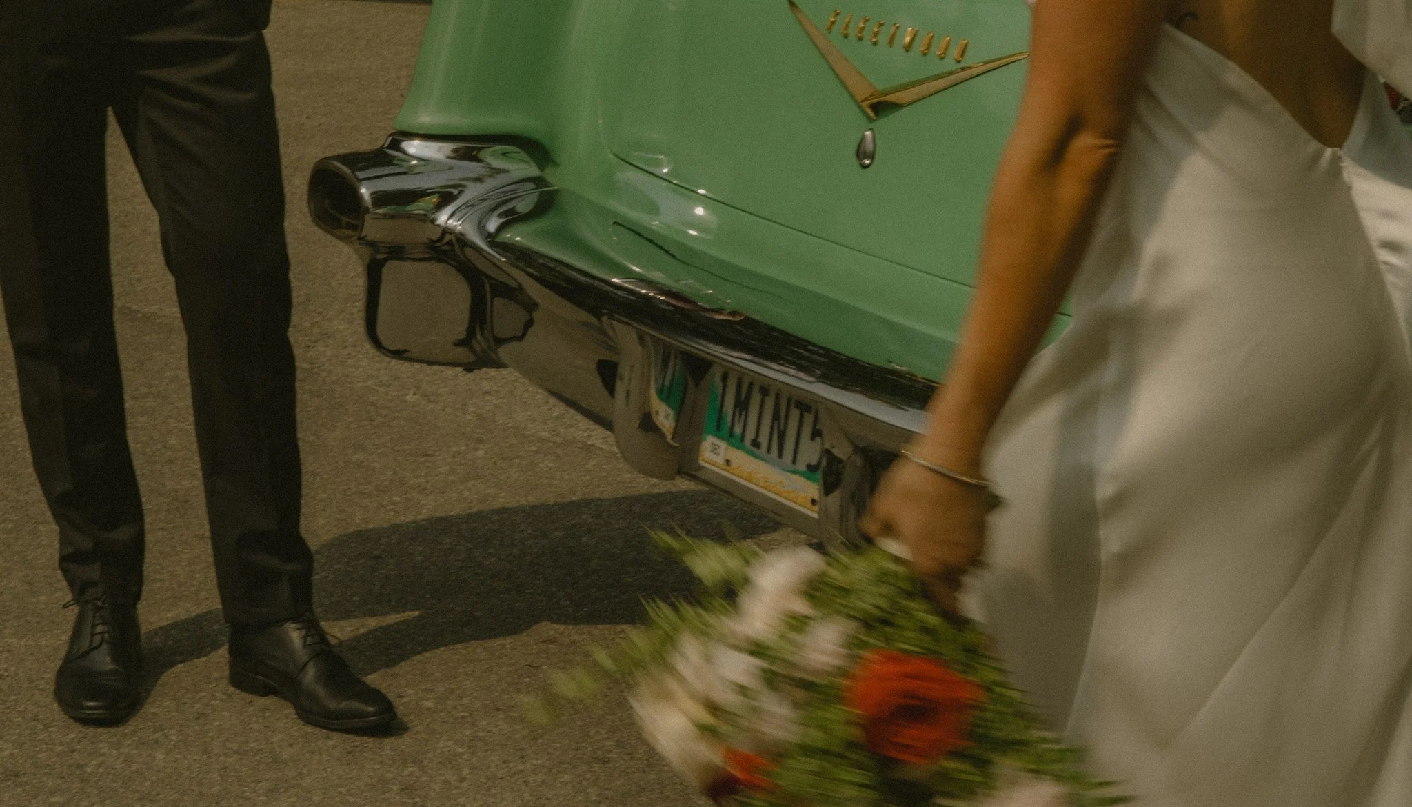 A person in a white dress holding a bouquet of flowers, standing next to a green vintage car with chrome details and a license plate reading '1 WINT3', accompanied by a man in black suit and shoes.