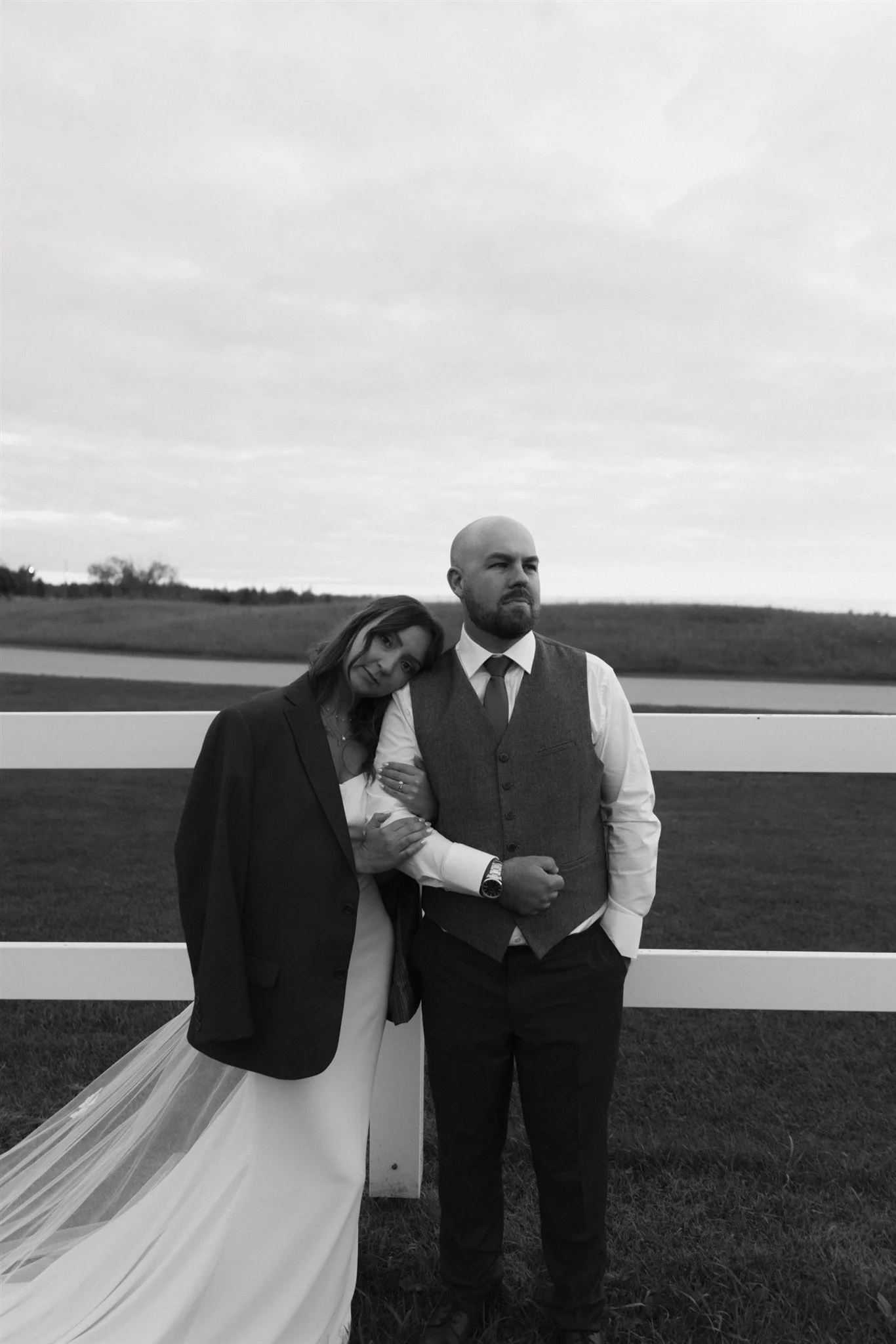 Black and white photo of a man and woman standing outdoors by a white fence. The woman leans her head on the man's shoulder, and they hold hands. The woman is wearing a wedding dress with a tuxedo jacket, and the man is dressed in a vest, shirt, and tie, with one hand in his pocket. The background shows an open field and a cloudy sky.