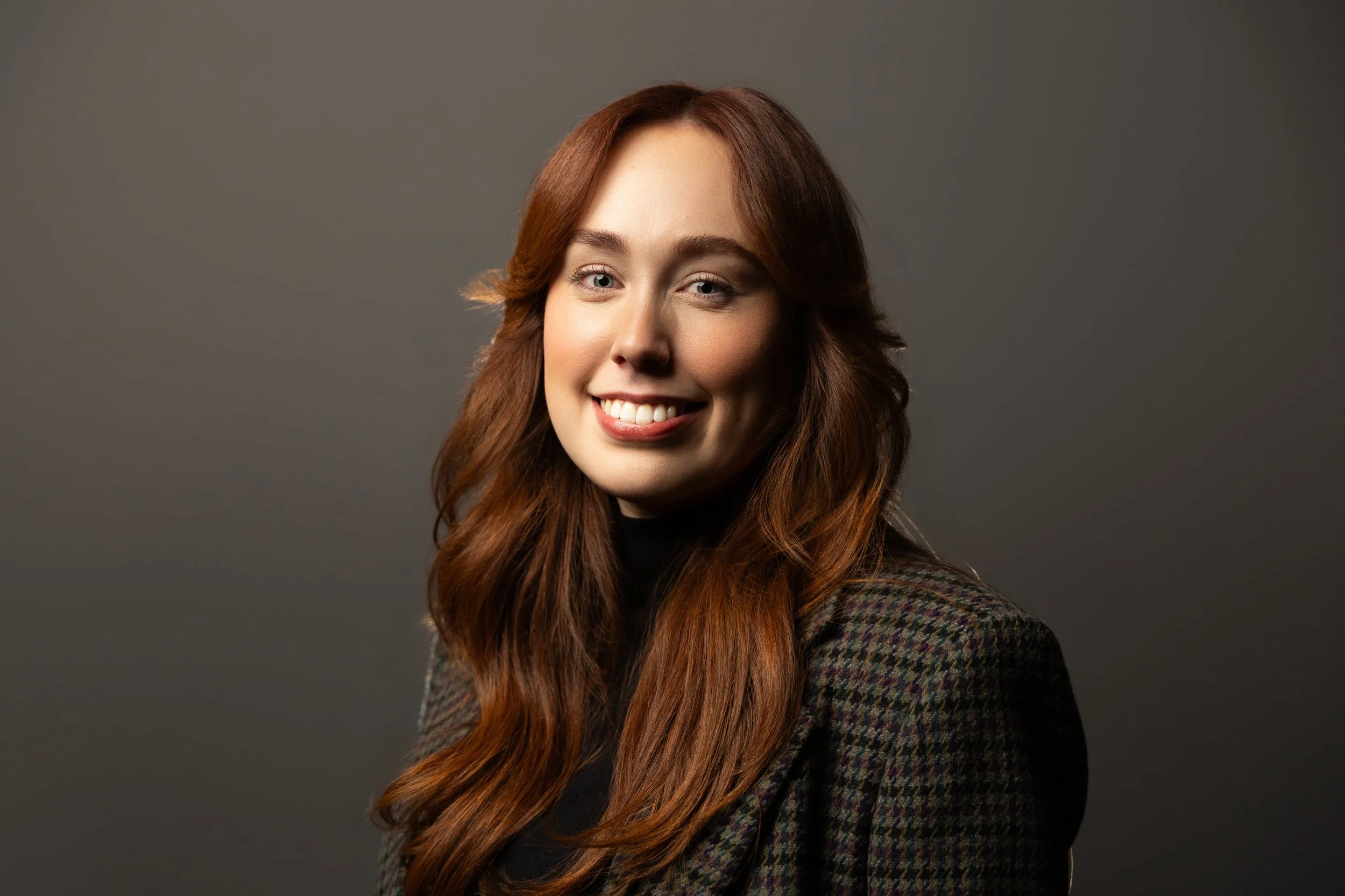 A striking woman with red hair and wearing a tweed blazer angles her head toward the camera and smiles for her headshot.