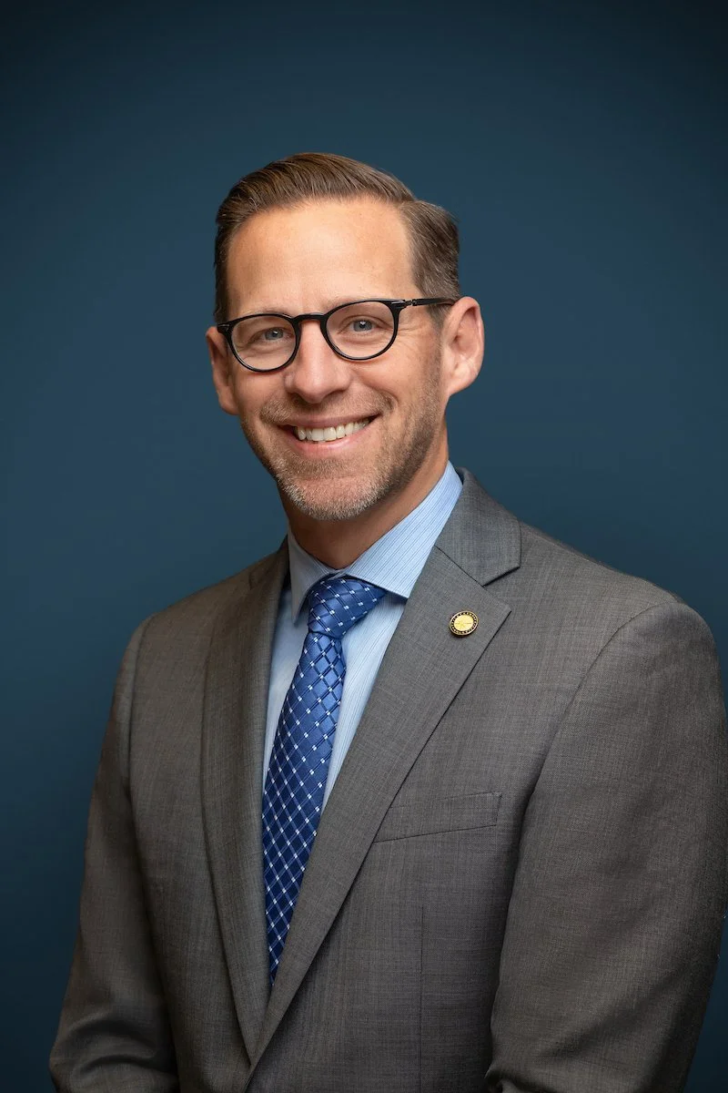 A man wearing a gray suit with a lapel pin and a blue tie poses against a blue backdrop matching his company brand color for his professional business portrait.