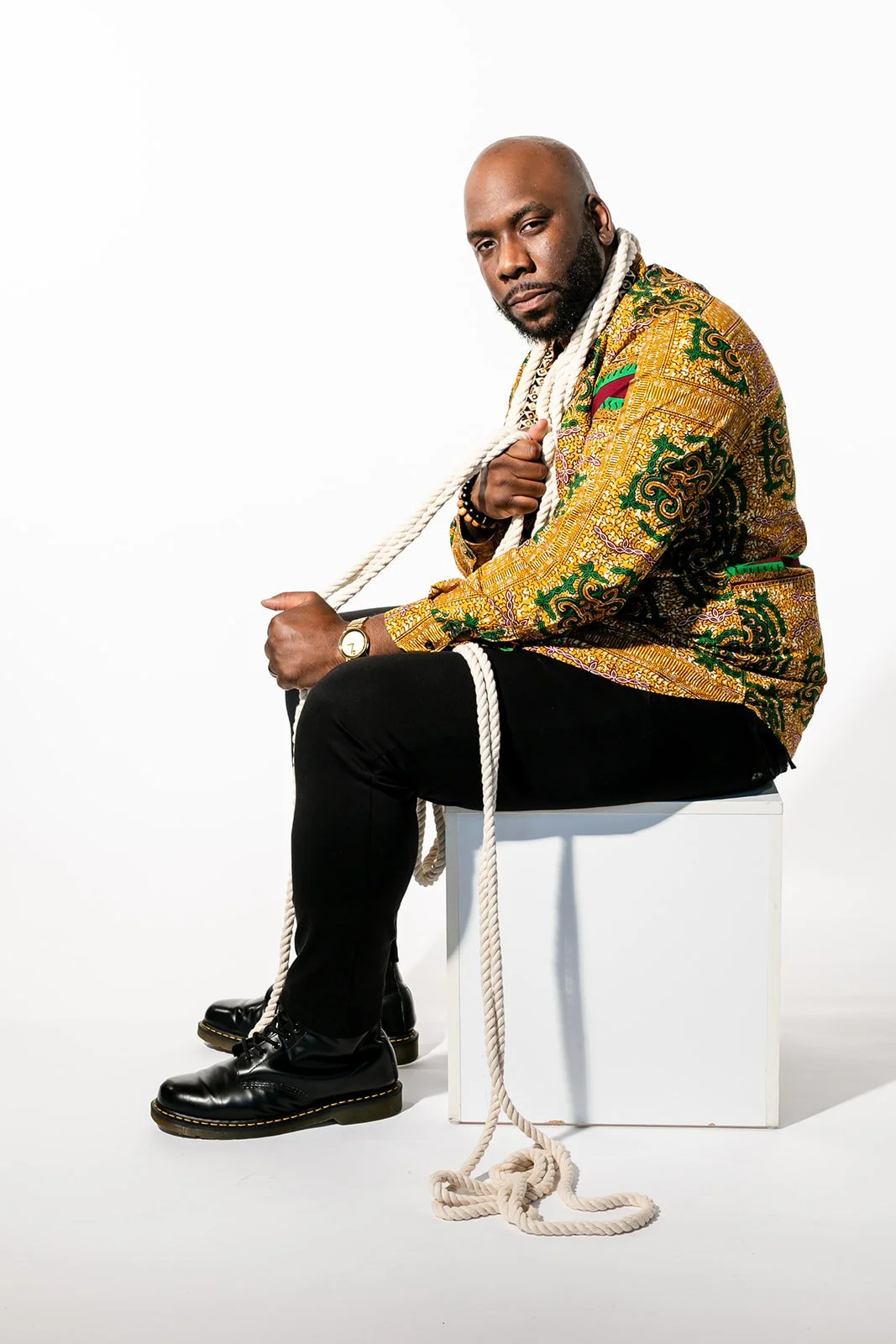 Black male Christian artist sitting on a white block, holding a thick white rope over his shoulder, wearing a colorful patterned jacket, black pants, and black boots against a plain white background.