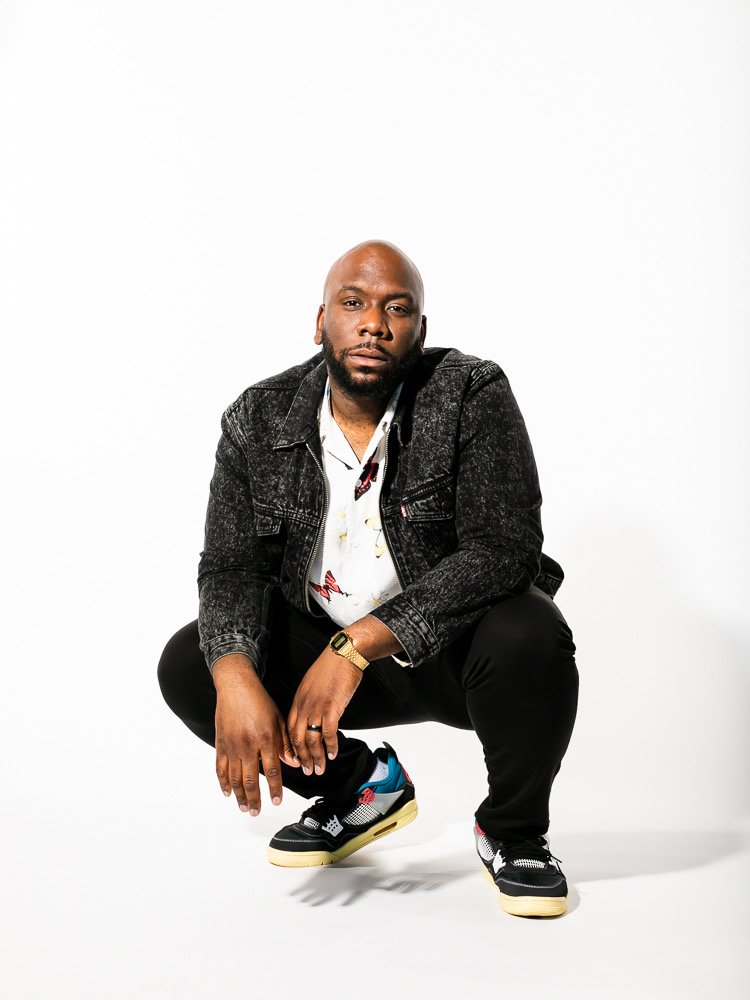 Man squatting in a studio with a white background, wearing a black denim jacket, white shirt, black pants, and colorful sneakers.