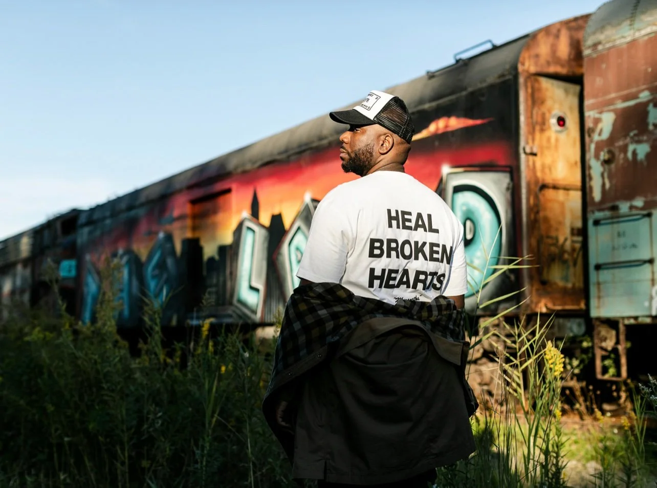 A black artist with a beard wearing a white T-shirt with the words 'Heal Broken Hearts' on the back, a cap, and a checkered shirt tied around his waist, standing outdoors near graffitied train cars with a sunset skyline painted on them.