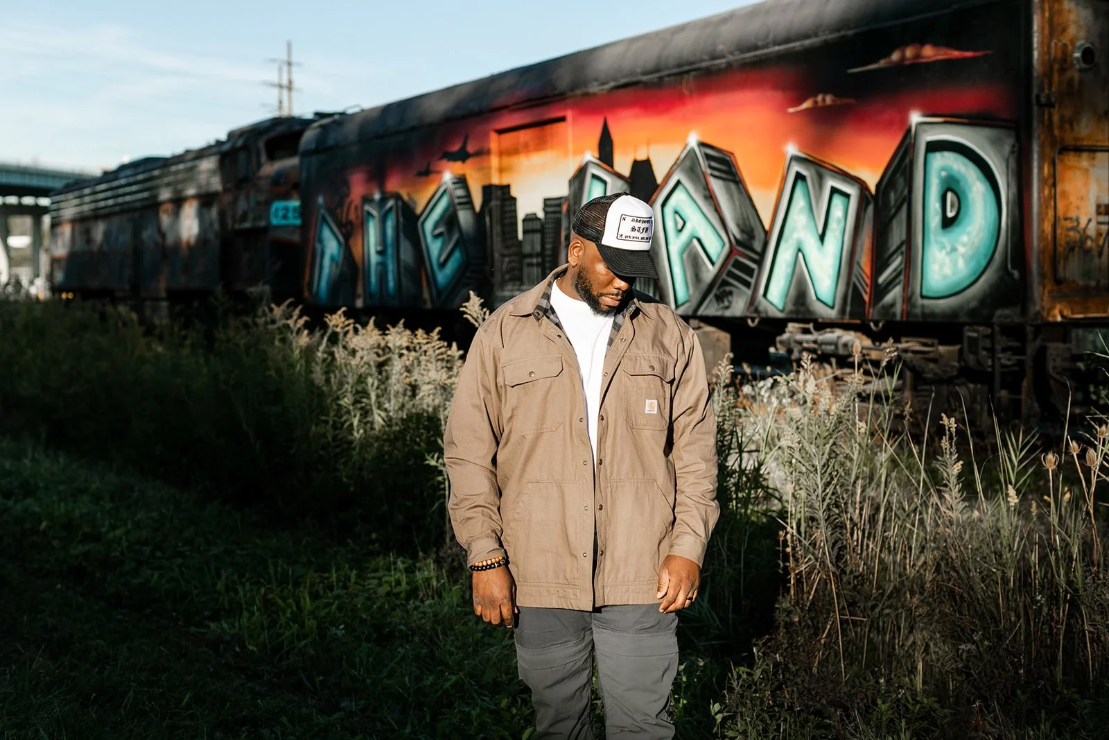 A man in a tan jacket and gray pants standing outdoors in front of an old train car with graffiti saying 'The Land' painted on it. The man is looking down, wearing a cap and beaded bracelet, with wild grass and plants around him.