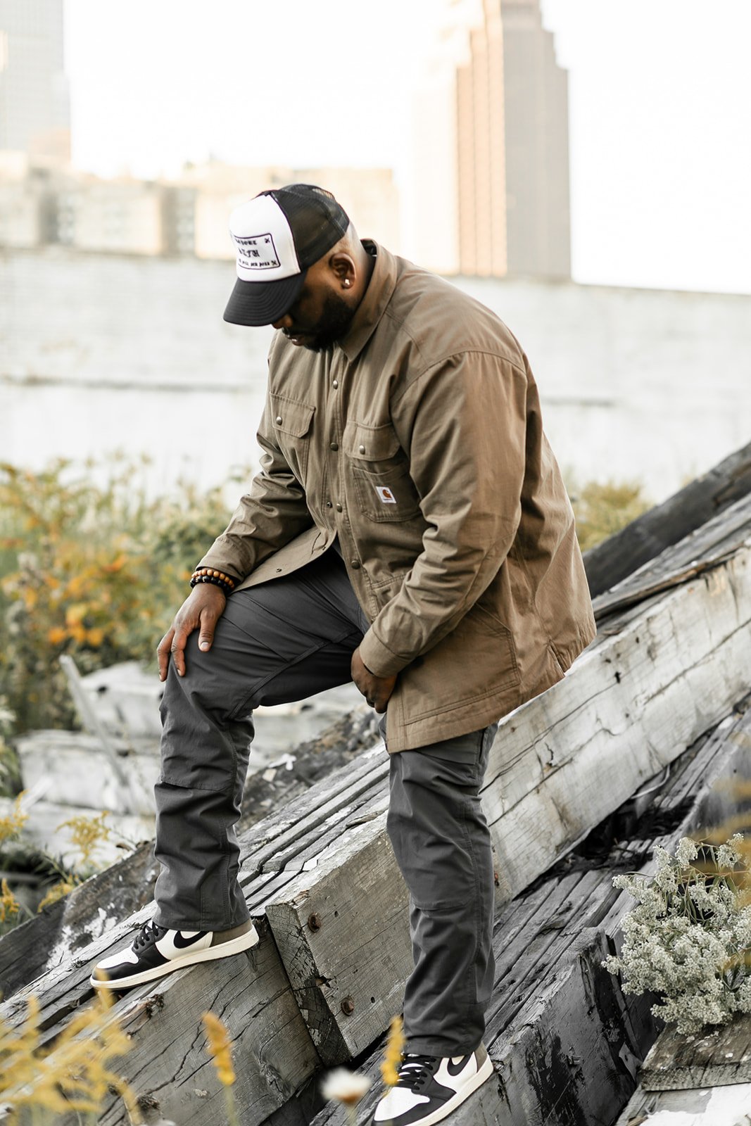 A man wearing a trucker hat, brown jacket, black athletic pants, and Nike sneakers standing on a wooden rooftop with city buildings in the background.