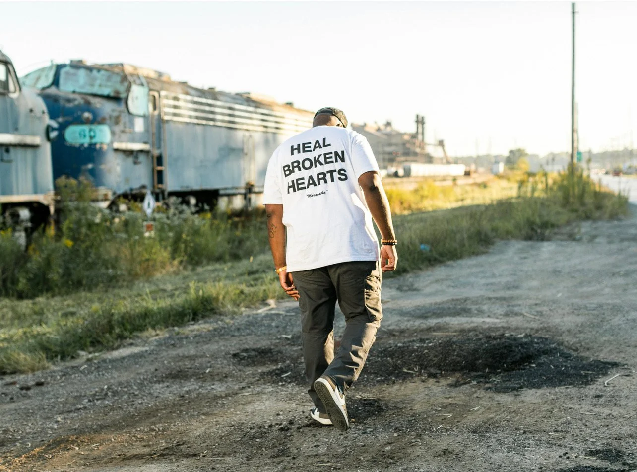 Man walking along a dirt path with abandoned train cars on the side, wearing a white T-shirt that says 'Heal Broken Hearts' on the back.