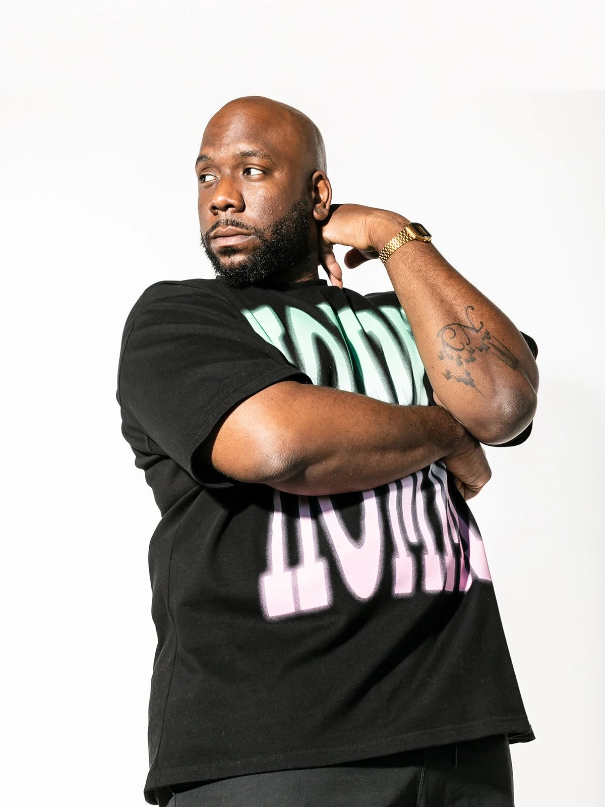 A man with a beard and a bald head, wearing a black T-shirt with colorful text, stands against a white background. He has tattoos on his right arm and wears a gold watch on his left wrist. He is looking to his left with his right hand behind his neck