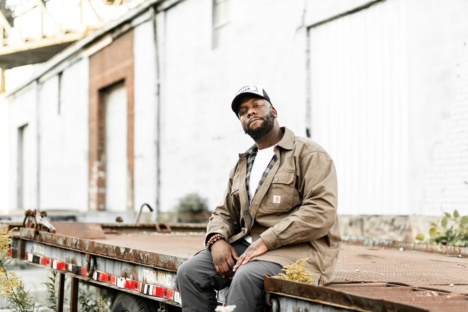 A man wearing a tan Carhartt jacket, gray pants, a black and white baseball cap, and beaded bracelets is sitting on the edge of a rusty flatbed truck in an urban setting with a white brick building in the background.