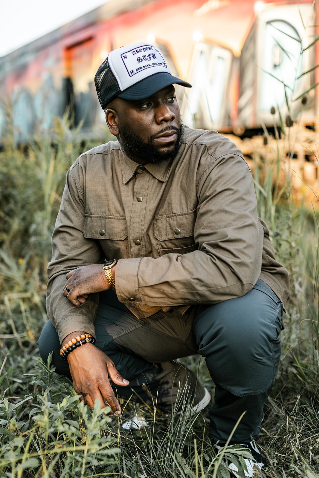 A man squatting outdoors near train tracks, wearing a beige jacket, black pants, and a trucker hat with writing on it. He is looking to the side with greenery around him.
