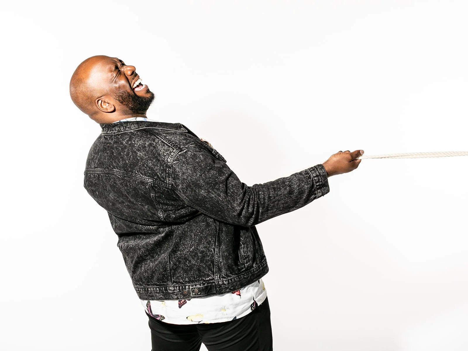 A man in a black, distressed denim jacket laughing while pulling on a rope.