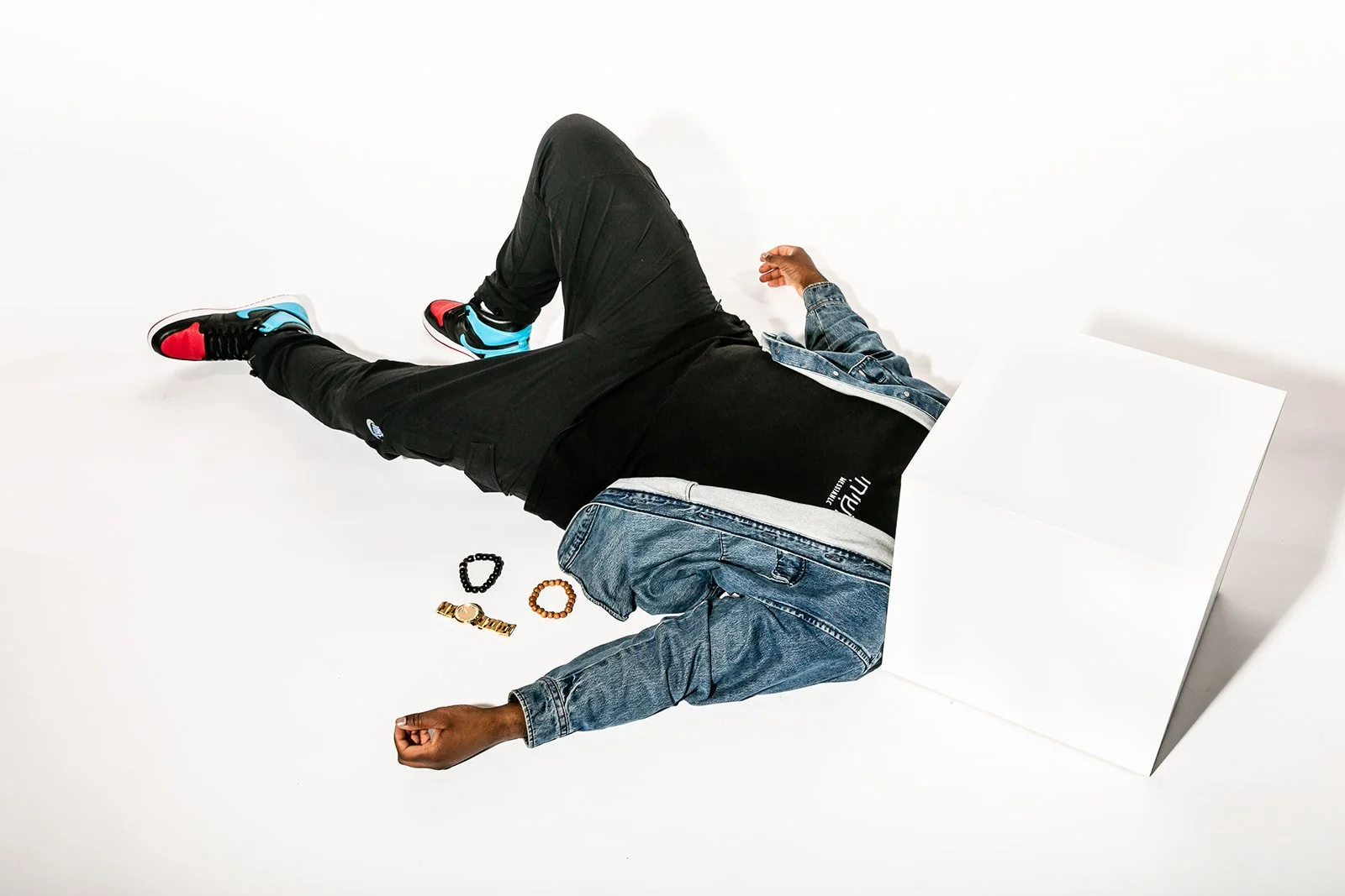 Person lying on the floor in black pants, black T-shirt, and jeans, with sneakers, with jewelry including a watch and bracelets, and a blank white sign leaning against a wall.