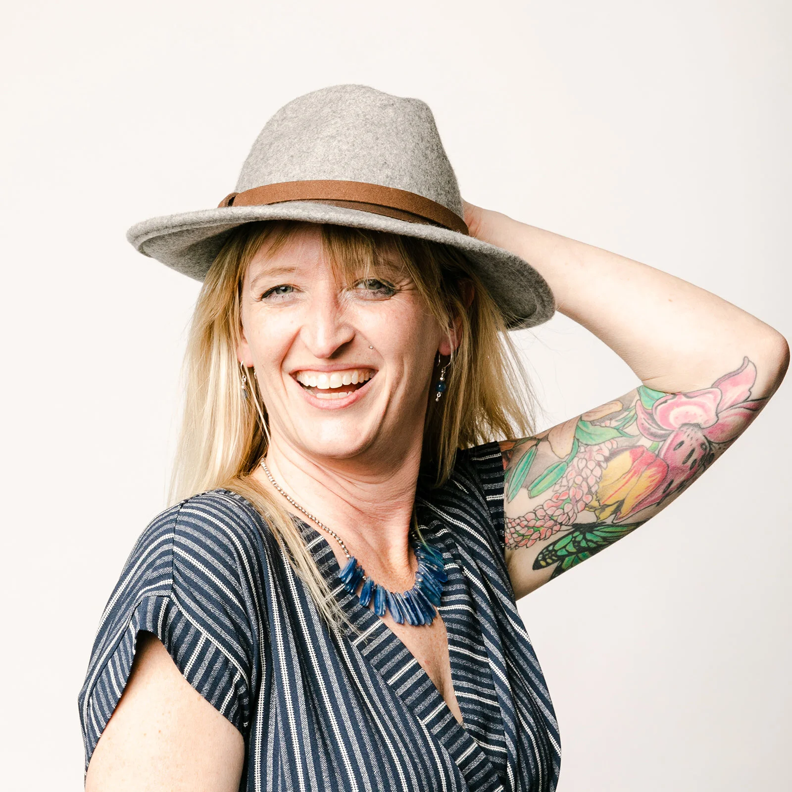 Woman with blonde hair holding a wool fedora on her head shows and her tattooed arm for her unconventional headshot 