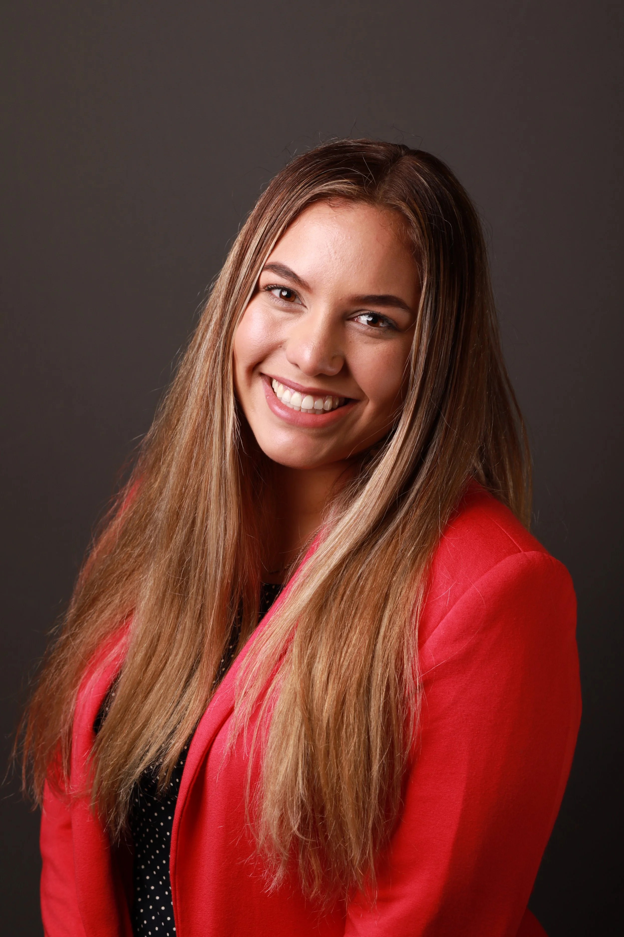 A young hispanic woman with long light brown hair, smiling and wearing a red blazer, in front of a dark gray background.