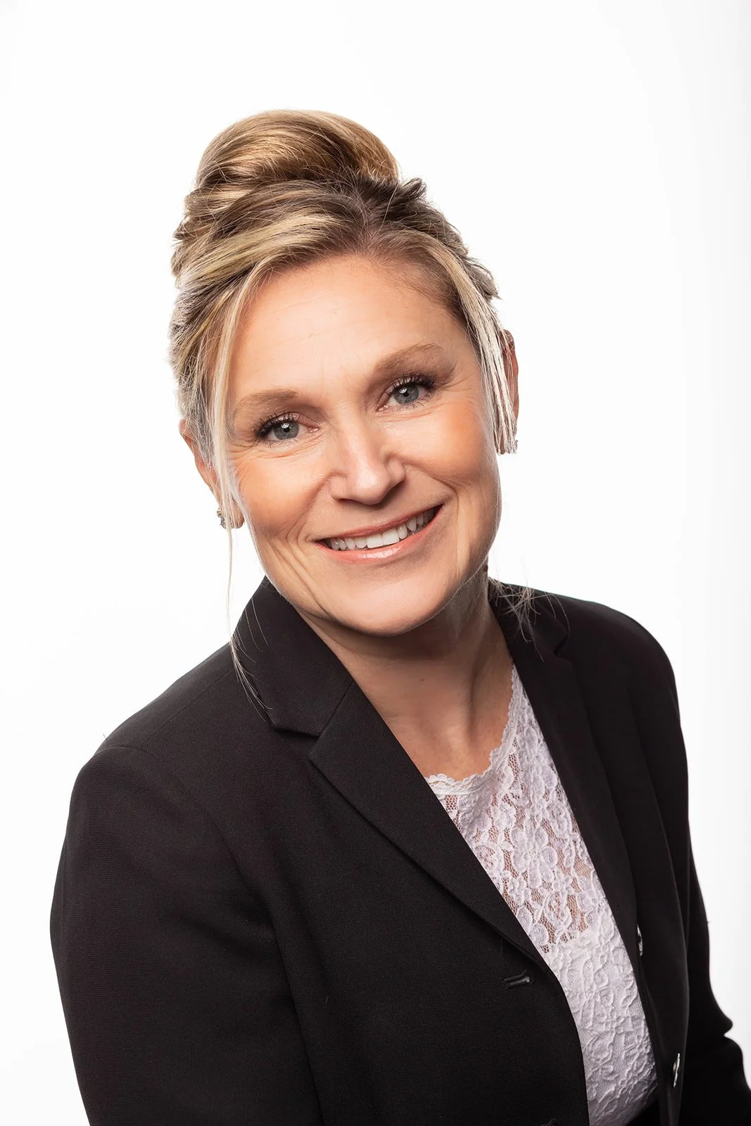 A professional woman with blonde hair styled in an elegant updo, wearing a black blazer and white lace top, smiling against a plain white background.