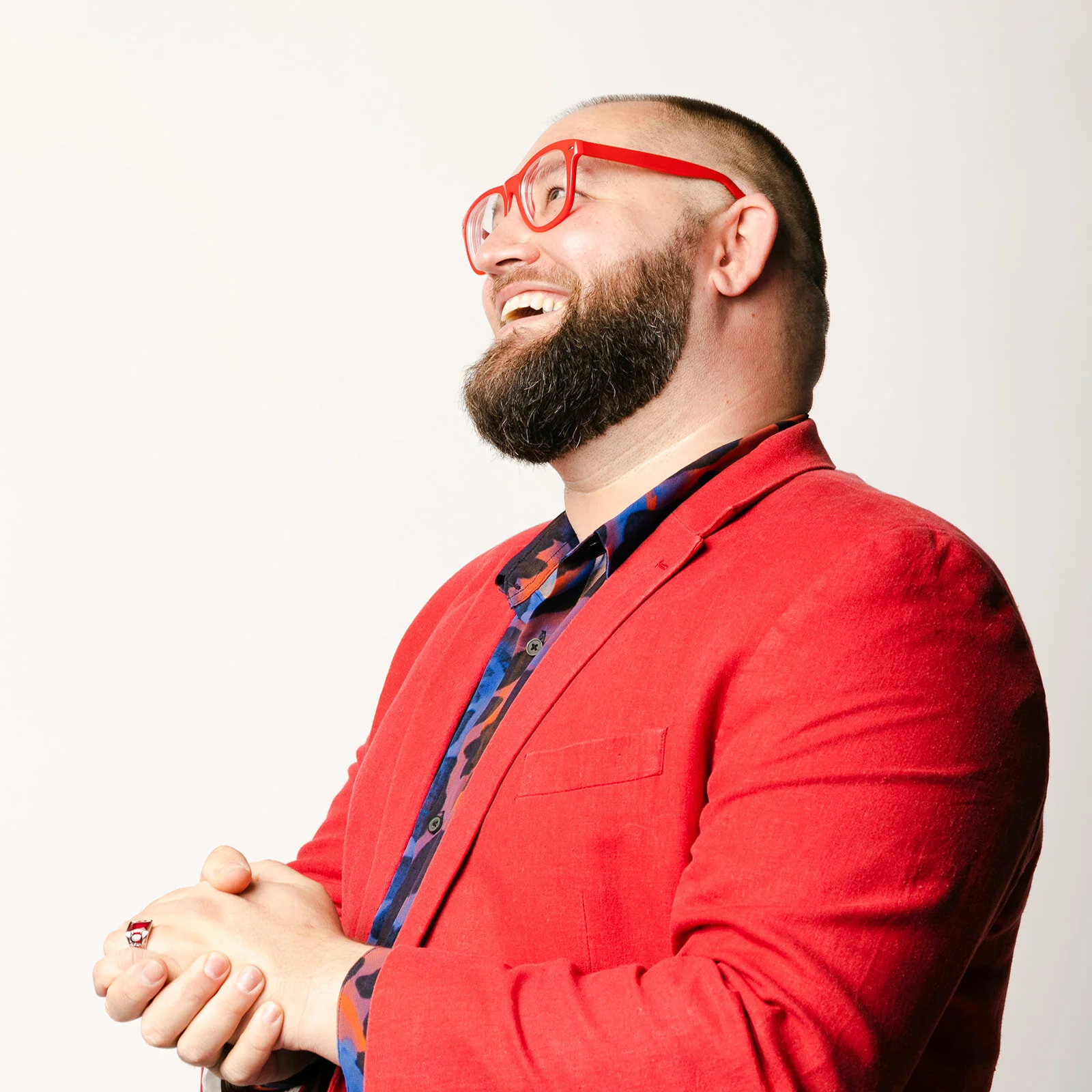 A smiling man with a beard, wearing red glasses, a red blazer, and a patterned shirt, standing against a plain background.