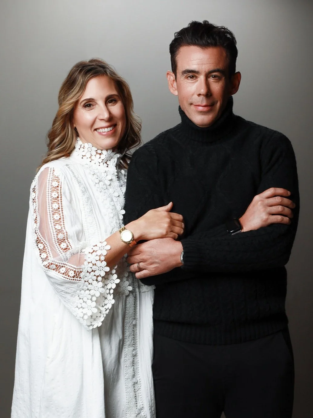 A smiling woman with wavy, shoulder-length blonde hair, wearing a white lace dress, stands to the left with her left hand on the arm of a serious-looking man with dark hair, wearing a black turtleneck sweater, standing to the right against a plain gr