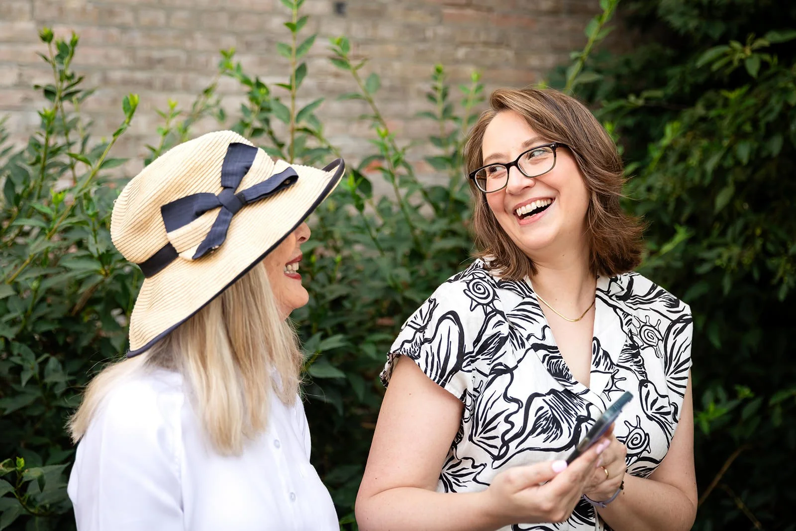 Two women outdoors, smiling and laughing, one holding a smartphone. One woman wears a wide-brimmed hat with a bow, the other wears glasses and a black-and-white patterned shirt.