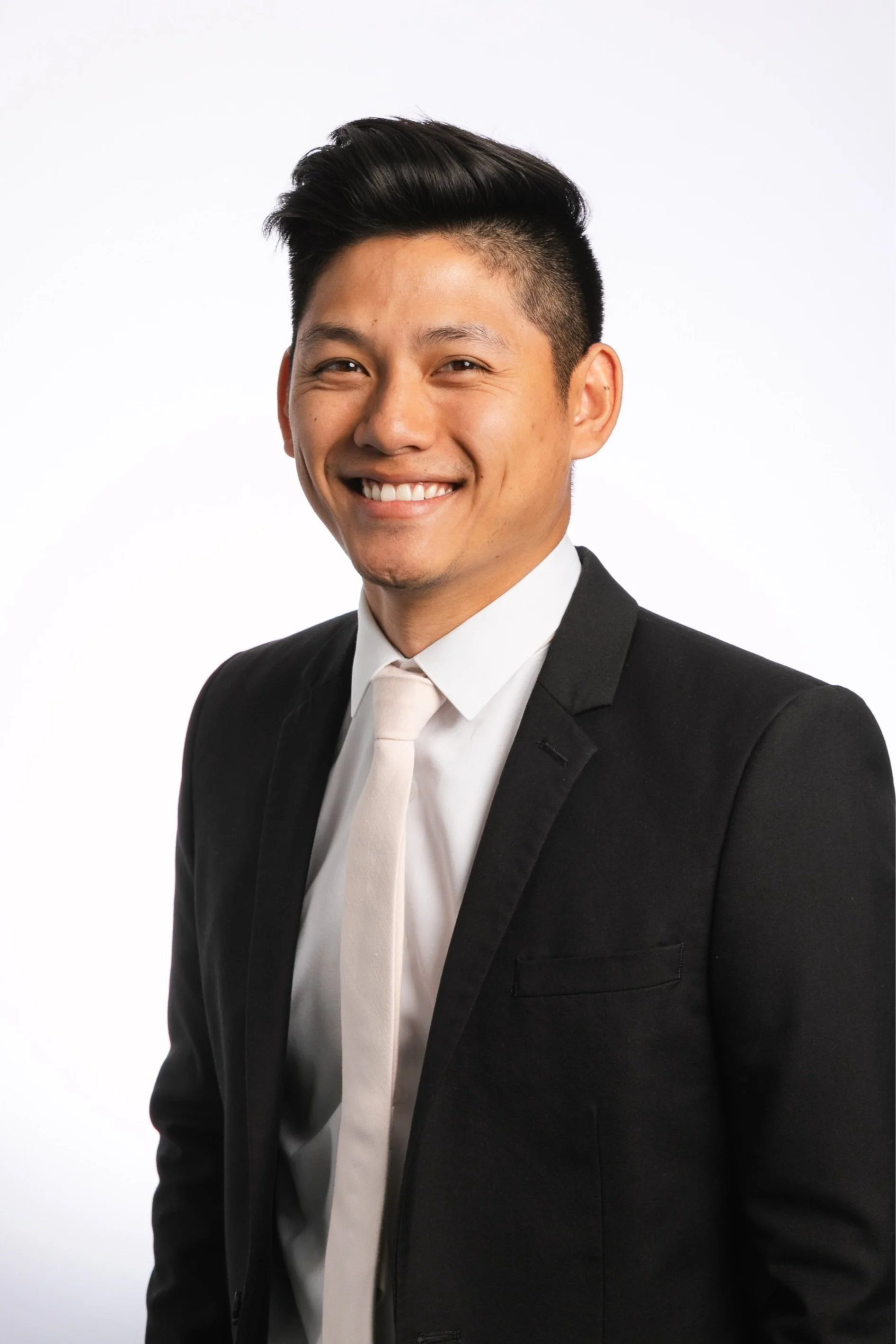 A smiling man dressed in a formal black suit with a white shirt and tie, posing against a plain white background.
