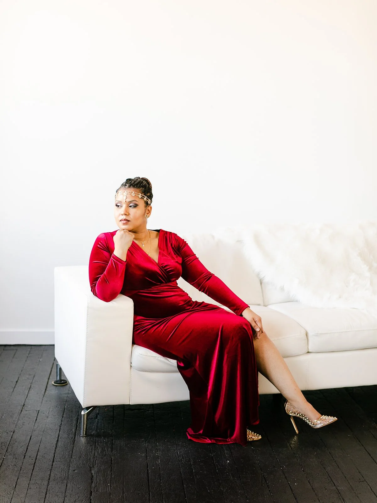 A woman in a long red velvet dress sitting on a white couch with her left arm resting on the armrest, her right hand touching her chin, and wearing silver heels and a headpiece, in a minimalistic room with white walls and dark wooden floors.