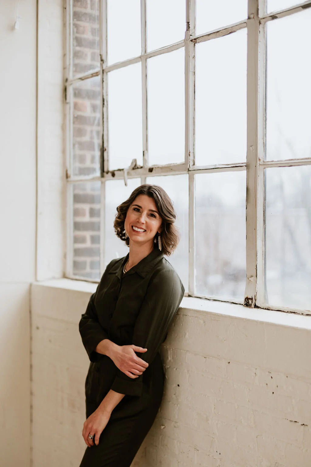 A woman with shoulder-length wavy hair, wearing a dark blouse, standing with arms crossed, smiling, next to a large industrial window with white frames and a brick wall visible outside.