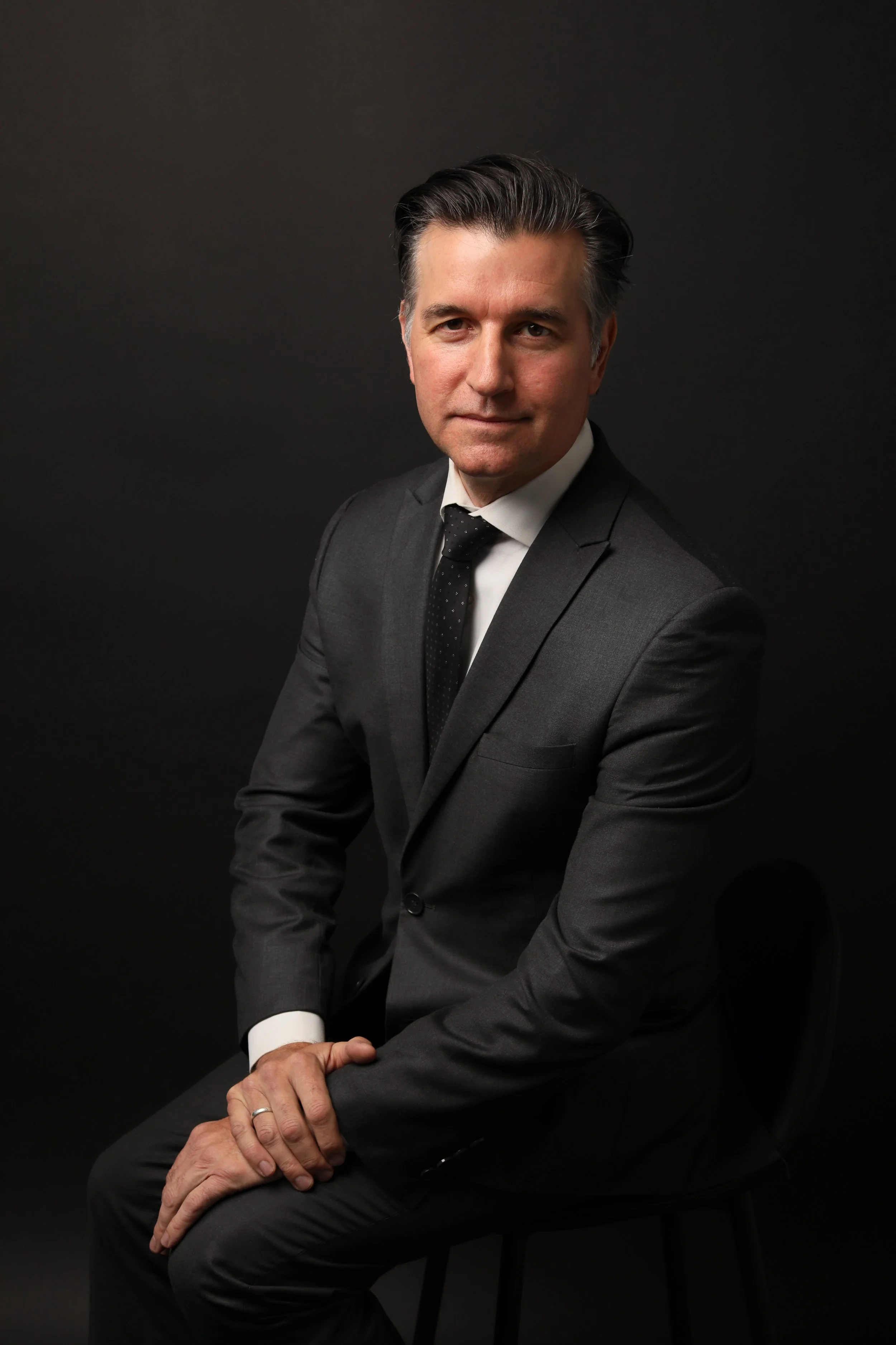 A business owner wearing a gray suit poses on a chair for his professional headshot in a dark, sophisticated background