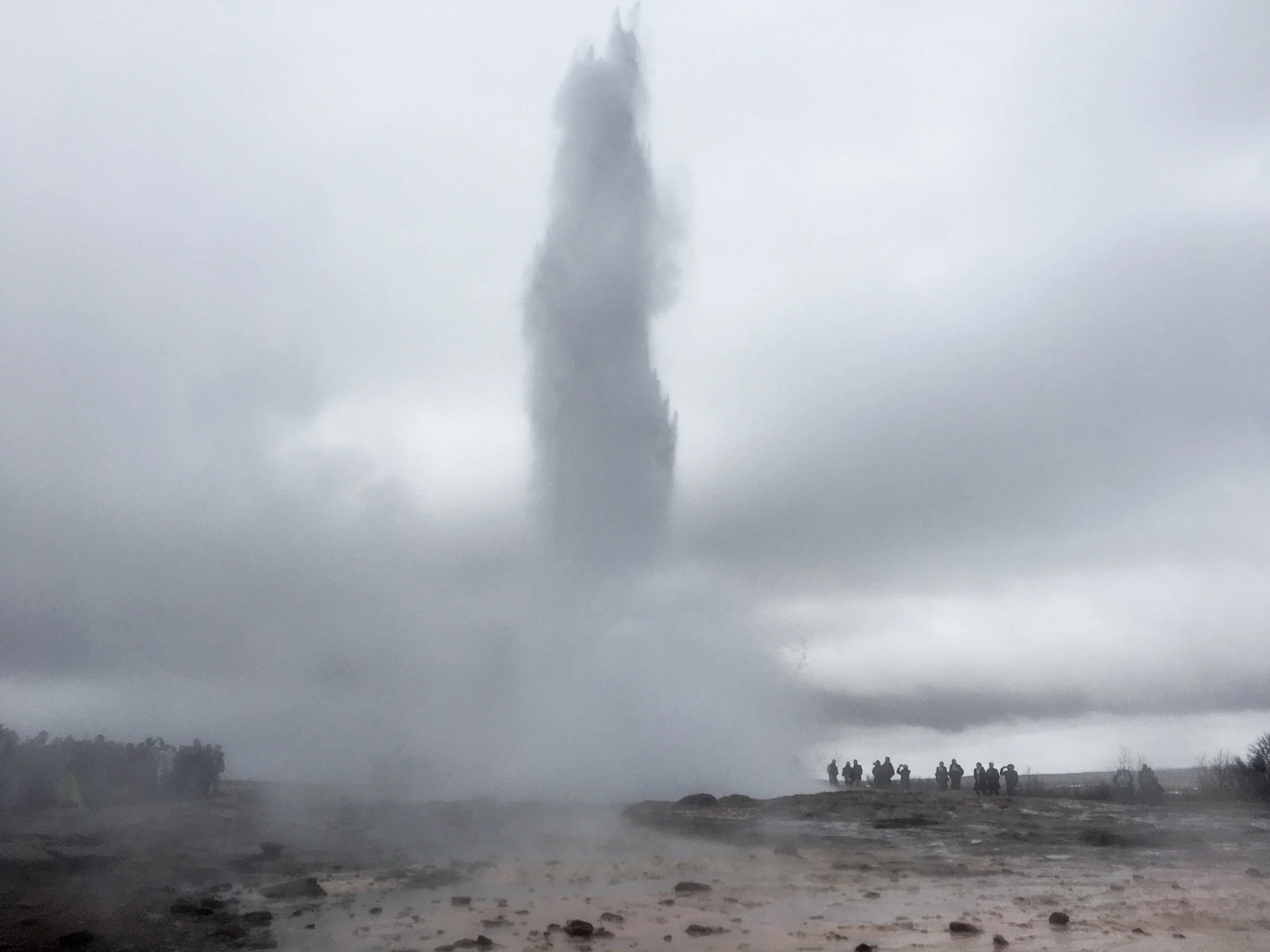 Large column of steam from a geyser in Iceland rising into cloudy sky, with a group of people and vehicles on a dirt road in the foreground.