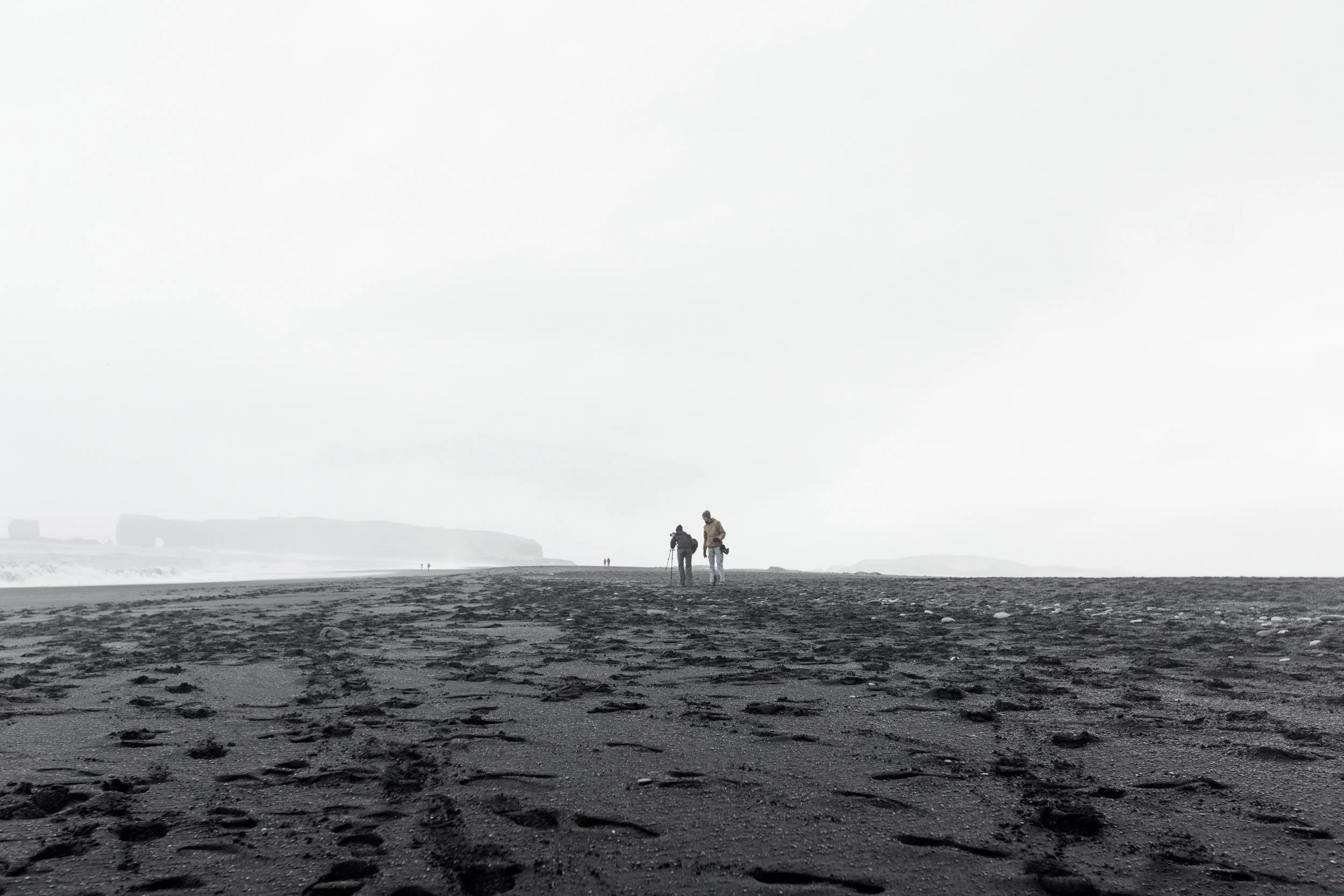 Two people walking on a black sandy beach with footprints, under a cloudy sky, with distant cliffs in the background.