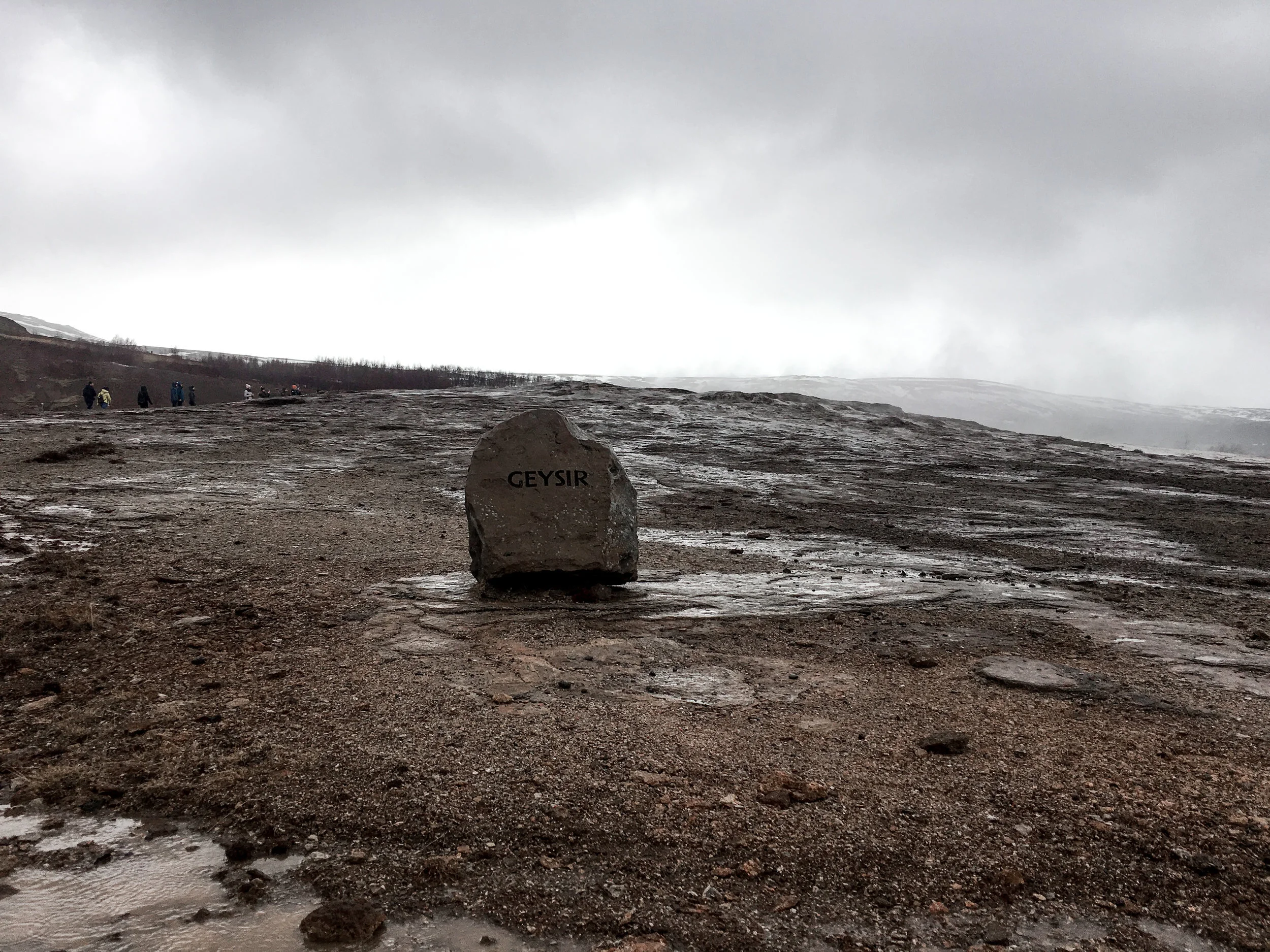 A rocky landscape with a large rock in the foreground labeled "Geysir" on a cloudy day, with a group of people in the distance and snow patches on the ground.