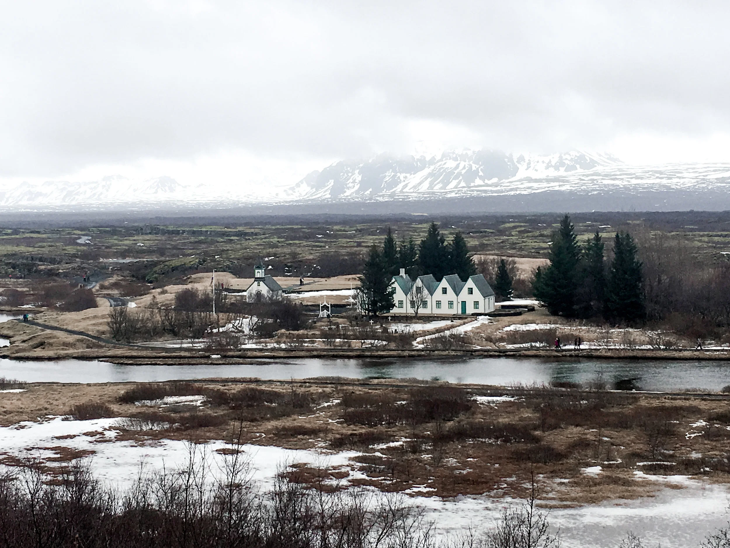 Scenic view of snow-capped mountains in the background, with a small village, white church, and several white houses in the middle ground, and a partially frozen river in the foreground, set in a cold, overcast environment.