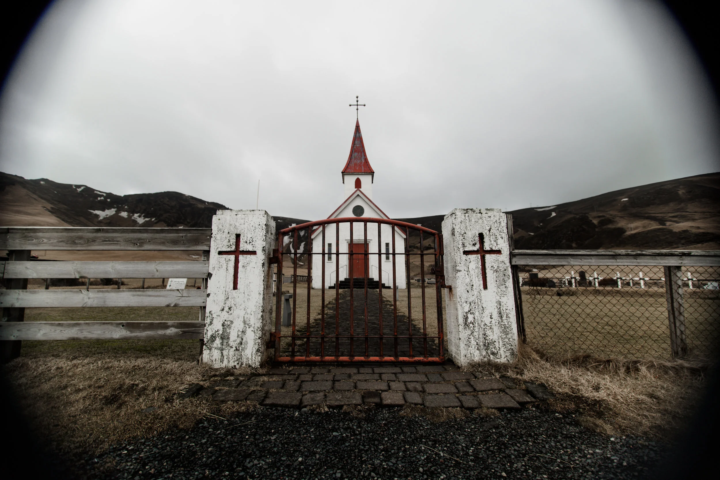 A white church with a red roof and a cross on top, behind a rusted gate with two red crosses on white pillars, surrounded by a field and hills under a cloudy sky.