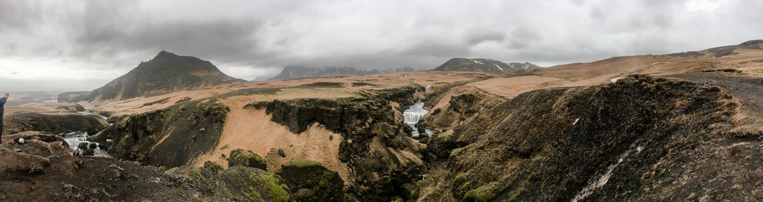 A rugged Icelandic landscape with moss-covered rocks, a small waterfall, rolling hills, and mountains under a cloudy sky.