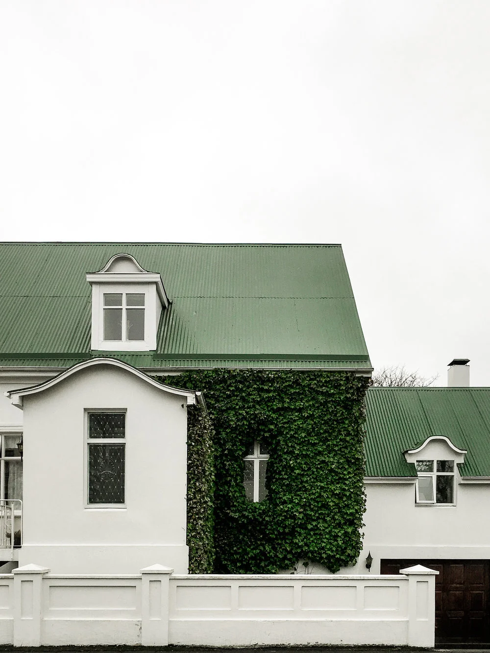 White house with green metal roof and ivy covering part of the exterior wall, windows with decorative panes, white fence