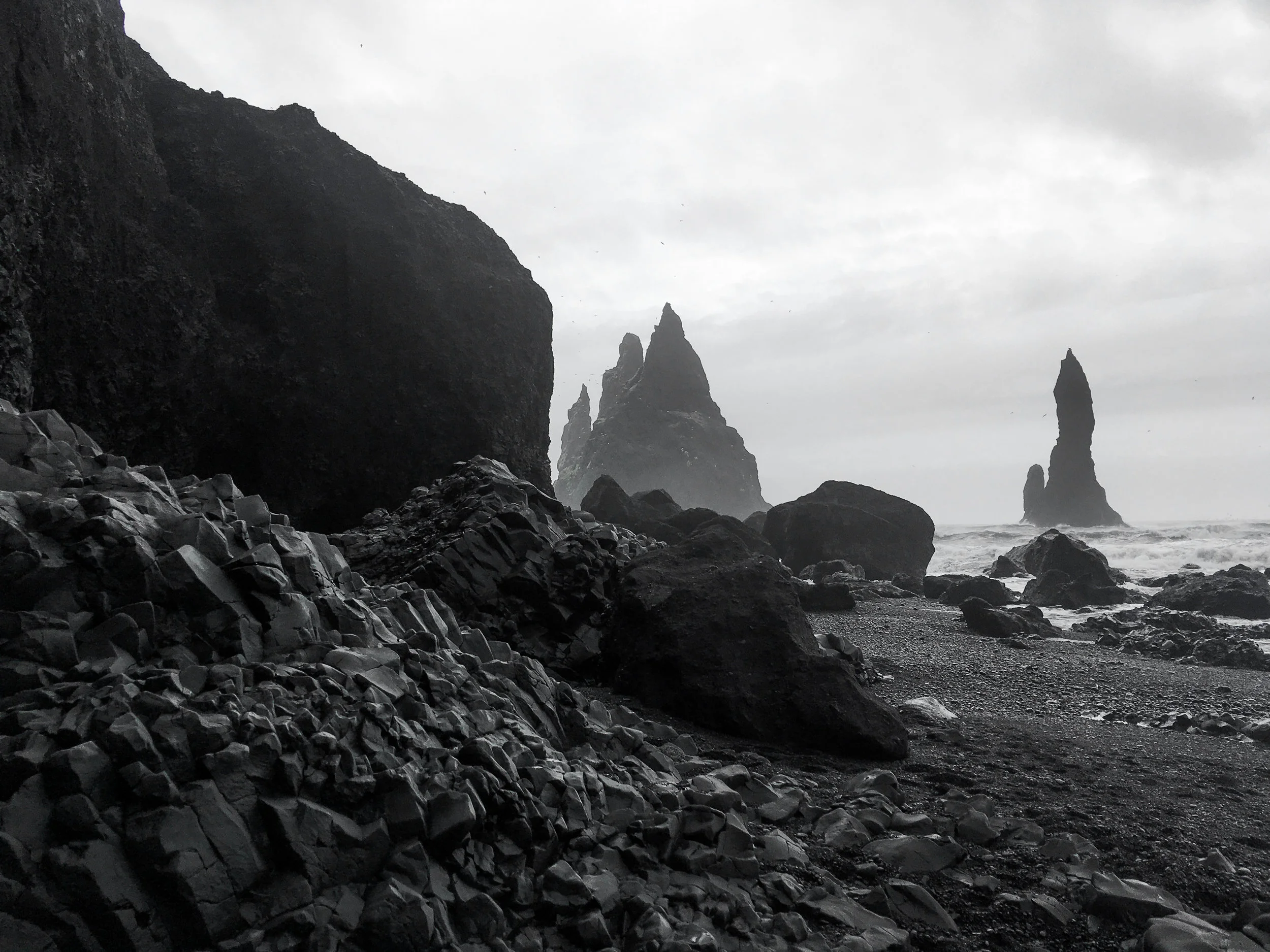 Black and white photograph of a rocky beach in Vik, Iceland, with large sea stacks in the ocean and clouds overhead.