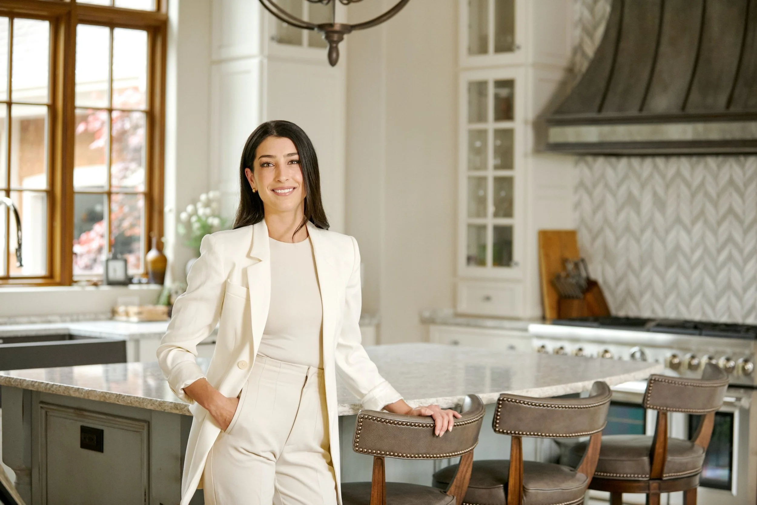 A real estate agent wearing an off-white business suit poses in a luxury, light kitchen for her commercial lifestyle photography session